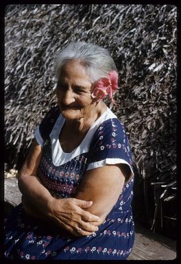 Elizabeth Paruai (Calico) Marsters, sitting next to a building with a thatched roof, Palmerston Island