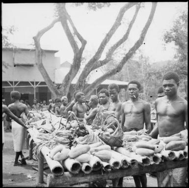 Men standing along one side of a laden trestle table, large tree behind, Boong, native market, Rabaul, New Guinea, ca. 1936 / Sarah Chinnery