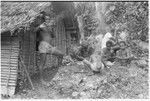 Taaboo lighting pipe in front of men's house with kids; another man in white shirt is pounding taro for pudding