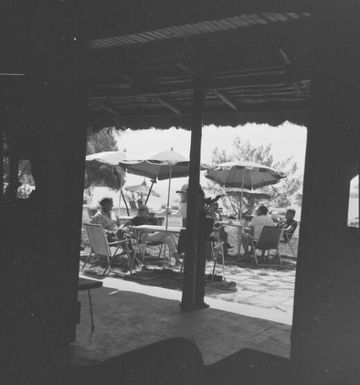 People sitting at tables with umbrellas outside a hut, Isle of Pines, New Caledonia, 1967 / Michael Terry