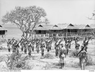 PAPUA, NEW GUINEA, 1942-07-20. "ALL PRESENT AND CORRECT, SIR". AN AUSTRALIAN INFANTRY PLATOON PARADES FOR RIFLE INSPECTION IN A PAPUAN COASTAL VILLAGE. THE STILTS OF THE NATIVE HUTS ARE IN THE ..