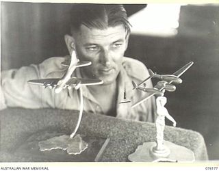 FINSCHHAFEN, NEW GUINEA. 1944-09-20. NX108519 LANCE CORPORAL A.P. MILLER, 18TH FIELD AMBULANCE, PROUDLY DISPLAYING TWO MODEL AIRCRAFT FASHIONED FROM SCRAP METAL