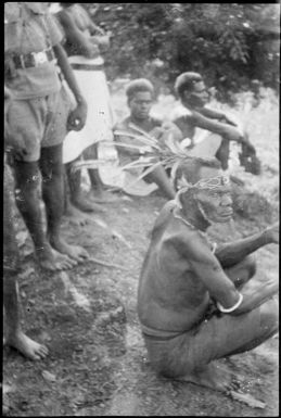 Seated man with feathers in his hair and a string design on his forehead with other people sitting and standing, New Guinea, ca. 1929 / Sarah Chinnery