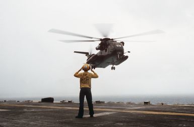 A plane director signals landing instructions to the pilot of a Helicopter Mine Countermeasures Squadron 14 (HM-14) RH-53D from the Sea Stallion helicopter hovering over the flight deck of the amphibious assault ship USS GUAM (LPH 9)