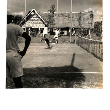 Photo of men playing tennis with a traditional hut in the background.