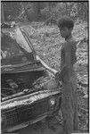 Truck with damaged front end, adolescent boy stands beside engine with improvised repairs