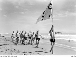 AITAPE, NORTH EAST NEW GUINEA. C. 1945-04. RAAF SURF PATROL DURING A MARCH PAST ALONG THE BEACH AT A SURF LIFESAVING CARNIVAL. STANDARD BEARER IS FLIGHT LIEUTENANT (FLT LT) BRUCE COTTRELL, SYDNEY, ..