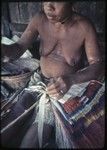 Weaving: woman makes skirt out of banana leaf fibers, some dyed red, blue or yellow, woman has word tattooed on her chest