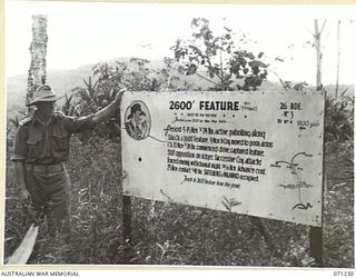 FINSCHHAFEN AREA, NEW GUINEA, 1944-03-17. VX5104 MAJOR V.J. SCHOFIELD, GENERAL STAFF OFFICER II, LIAISON OFFICER OF THE 2ND AUSTRALIAN CORPS, PICTURED ALONGSIDE A BATTLE SIGN AT THE 2,600 FEET ..