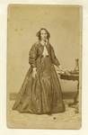 Studio portrait of an unidentified woman, whole-length, standing, to left, resting hand on book on table