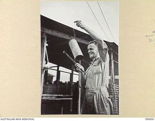 CAPE WOM, WEWAK AREA, NEW GUINEA. 1945-09-15. SERGEANT J. HANSON, HEADQUARTERS 6 DIVISION, RINGING THE MESS GONG. THE GONG IS MADE FROM AN OLD USED CARTRIDGE CASE OF A 25-POUNDER SHELL