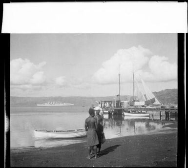 Two men standing beside a beached rowboat nd a jetty with moored ships and HMAS Australia [?],   Rabaul Harbour, New Guinea, 1937 / Sarah Chinnery