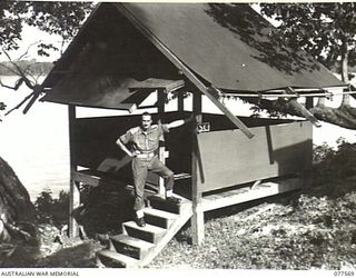 NX70682 CAPTAIN A. MCC. HEATHWOOD, STAFF CAPTAIN, HEADQUARTERS, 23RD INFANTRY BRIGADE STANDING ON THE STEPS OF HIS NEW OFFICE BUILDING