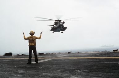 A plane director signals landing instructions to the pilot of a Helicopter Mine Countermeasures Squadron 14 (HM-14) RH-53D Sea Stallion helicopter hovering over the flight deck of the amphibious assault ship USS GUAM (LPH 9)