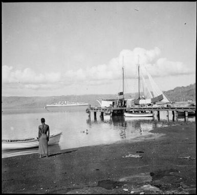 Man standing beside a beached rowboat and a jetty with moored ships and HMAS Australia [?] in the background, Rabaul Harbour, New Guinea, 1937 / Sarah Chinnery