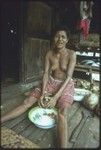 Food preparation: woman shreds coconut meat into bowl, more coconuts, yams and other foods in background