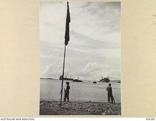 MALAGUNA MISSION, NEW BRITAIN. 1945-09-10. TROOPS OF 4 INFANTRY BRIGADE ON THE BEACH. THE AUSTRALIAN FLAG IS IN FOREGROUND AND SHIPPING IN SIMPSON HARBOUR IN BACKGROUND. THE TROOPS NOW OCCUPY THE ..