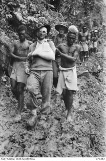 N130859 Private (Pte) E J Pinney, 9th Infantry Battalion (front, with his arm in a sling) being assisted down the track from George Hill by two natives. Pte Pinney was wounded by a mortar bomb ..