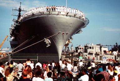 Family and friends watch from the pier as crew members man the rails aboard the amphibious assault ship USS GUAM (LPH-9). The large harbor tug WATHENA (YTB-825) and another tug are maneuvering the GUAM as the vessel prepares to depart for the Middle East in support of Operation Desert Shield