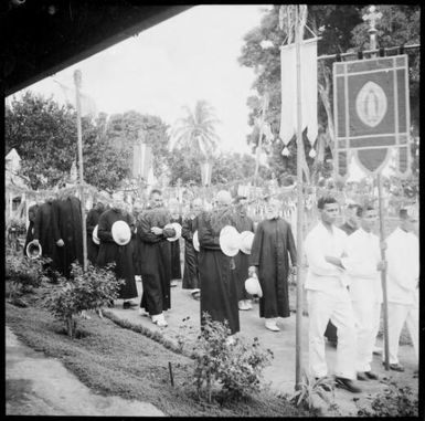 Man holding a banner with two other men leading a group of priests in the Corpus Christi Procession, Vunapope Sacred Heart Mission, Kokopo, New Guinea, 1937 / Sarah Chinnery