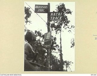 BOUGAINVILLE. 1945-04-22. LANCE-CORPORAL E.A. DUKE, 24 INFANTRY BATTALION, LOOKING AT A SIGN AT ANDERSON'S JUNCTION, WHICH WAS NAMED AFTER THE COMMANDING OFFICER OF THE UNIT. THE LOWER NOTICE WARNS ..