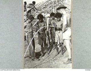 PORT MORESBY, PAPUA. 1942-07-20. PAPUAN NATIVES ARE EMPLOYED IN ERECTING GRASS AND WOOD HUTS FOR AMERICAN TROOPS. HERE TWO AMERICAN SOLDIERS ARE SEEN WATCHING NATIVE "BOYS" STRIPPING THE BARK