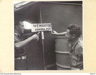 KARAWOP, NEW GUINEA, 1945-09-18. CAPTAIN J.R. THOMPSON (1) AND PRIVATE L.J. CLEARY (2), 2/6 CAVALRY COMMANDO REGIMENT, STANDING BESIDE THEIR DENTAL POST SIGN