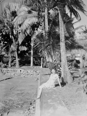 [Portrait of a woman sitting on a wall by palms]