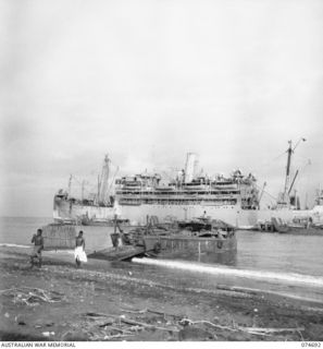 LAE, NEW GUINEA. 1944-07-13. NATIVES UNLOADING SAC-SAC (SAGO PALM ROOFING THATCH) AT THE HEADQUARTERS NEW GUINEA FORCE WHARF. THE AUSTRALIAN VESSEL HMT "ORMISTON" IS ANCHORED IN THE BACKGROUND