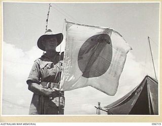 CAPE WOM, WEWAK AREA, NEW GUINEA, 1945-09-20. PRIVATE S. DAVIS, 2/4 INFANTRY BATTALION WITH HIS FAMOUS JAPANESE FLAG. IN THE RED SPOT, CENTRE OF FLAG, ARE THE NAMES OF MEMBERS OF C COMPANY, 2/4 ..