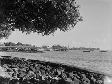 [View of a pier and boats anchored off-shore as seen from a stone-lined beach]