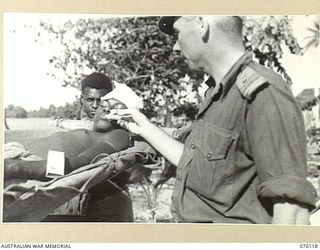 MILILAT, NEW GUINEA. 1944-09-21. MR. R.G. SCOTT, AUSTRALIAN RED CROSS SOCIETY ATTACHED TO HEADQUARTERS 5TH DIVISION, PASSES A CIGARETTE TO A WOUNDED NATIVE ARRIVING AT THE AUSTRALIAN NEW GUINEA ..