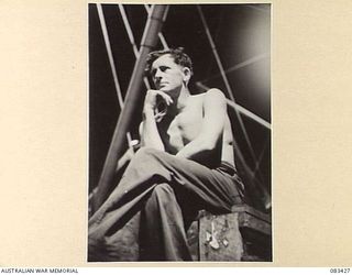 FINSCHHAFEN, NEW GUINEA, 1944-11-15. SAPPER N.A. SANDERSON, 2/8 FIELD COMPANY, ROYAL AUSTRALIAN ENGINEERS, ILLUMINATED BY DECK LAMPS ABOARD USS MEXICO, LOOKS TOWARDS SHORE LIGHTS AND THE SCENE OF ..
