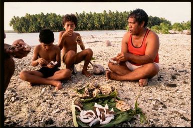 Two boys and a man eating a meal on beach, Cook Islands