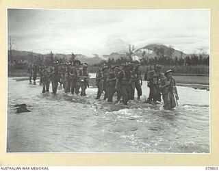 BOUGAINVILLE, SOLOMON ISLANDS. 1945-03-19. TROOPS OF THE 58/59TH INFANTRY BATTALION CARRYING FULL PACKS, CROSS THE TOROKINA RIVER. MOUNT BAGANA, AN ACTIVE VOLCANO, LIES IN THE DISTANCE