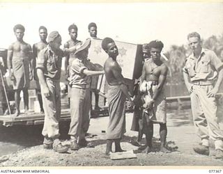 NEW GUINEA. 1944-12-14. NATIVES CARRYING CASES OF AUSTRALIAN COMFORTS FUND GOODS ASHORE FROM A LANDING BARGE