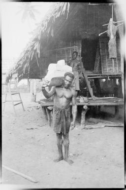 Man with a load on his shoulder and a house behind him, Awar [?], Sepik River, New Guinea, 1935 / Sarah Chinnery