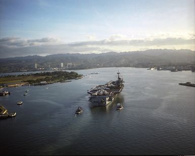 An aerial stern view of the US Navy (USN) Forrestal Class Aircraft Carrier USS RANGER (CV 61), with her Sailors manning the rails and aircraft of the Carrier Air Wing 2 (CVW-2) on her deck, trailed by tugboats, as she arrives at Pearl Harbor Naval Base in Pearl Harbor, Hawaii (HI). To the left are the USS ARIZONA (BB 39) Memorial and Ford Island