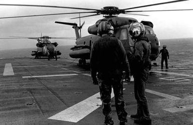 Two crewmen discuss their duties on the flight deck of the amphibious assault ship USS GUAM (LPH 9), as two CH-53 Sea Stallion helicopters are prepared for takeoff during operations off the coast of Beirut, Lebanon