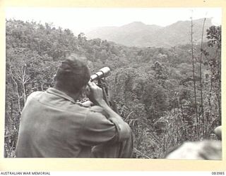 TOROKINA, BOUGAINVILLE, SOLOMON ISLANDS. 1944-12-15. SIGNALMAN F.C. CHRISTENSEN, 9 INFANTRY BATTALION, AT GEORGE HILL, OBSERVING WITH A TELESCOPE, MT. BAGANA, AN ACTIVE VOLCANO STANDS AT THE ..