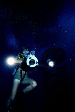 Underwater view of a Navy photographer wearing scuba gear with an American flag in the background attached to the hull of submarine ex-USS BLUEGILL (SS 242) during Pacific Submarine Salvage Exercise 83 (PACSUBSALVEX-83) off the coast of Maui