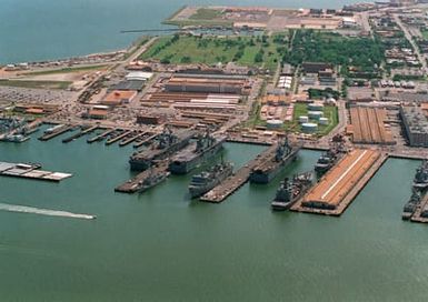 An aerial view looking northeast, of a section of the base showing various supply storage buildings and fuel storage tanks. In the lower section of the photograph can be seen the amphibious assault ship USS WASP (LHD-1) tied up on the north side of pier 7 with the USS SAIPAN (LHA-2) tied up on the south side. The USS NASSAU (LHA-4) is tied up on the south side of pier 6