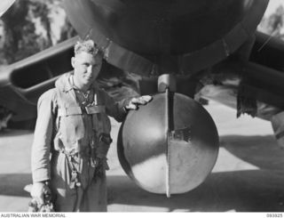 PIVA AIRSTRIP, TOROKINA, BOUGAINVILLE, 1945-07-12. SQUADRON LEADER W.A. HARDHAM, ROYAL NEW ZEALAND AIR FORCE, ALONGSIDE LIEUTENANT KINNA'S MOVIE CAMERA WHICH HAS BEEN FITTED INTO THE BELLY TANK OF ..