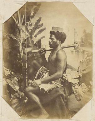 Kanak man seated posing with native implements against studio backdrop, New Caledonia, ca. 1870s / Allan Hughan