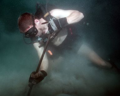 Aviation Ordinanceman First Class (AO1) Jonathan A. Hamm, Explosive Ordnance Disposal Mobile Unit Five (EODMU5), Detachment Marianas, uses a pry bar to unearth Hedge Hog Rockets, 105mm, 5 inch 50, 20mm, and 50 caliber rounds from a barge that sunk after WWII