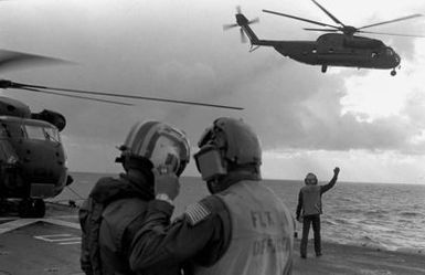 Two crewmen discuss their duties on the flight deck of the amphibious assault ship USS GUAM (LPH 9), as a CH-53 Sea Stallion helicopter takes off during operations off the coast of Beirut, Lebanon