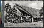 Men stand under the opening of a large thatched pavilion, Papua New Guinea, ca.1900-1930