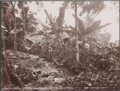 Two men standing next to a stone wall that surrounds a village at Te Motu, 1906 / J.W. Beattie