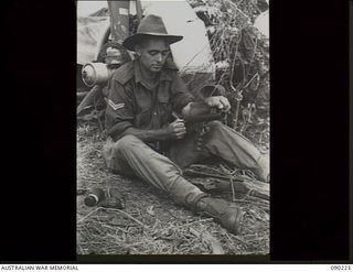 DAGUA, NEW GUINEA. 1945-03-30. CORPORAL S.G. HARTLEY, 2/2 INFANTRY BATTALION, SITTING NEAR A JAPANESE AIRCRAFT ENGINE ON THE AIRSTRIP, CLEANING HIS OWEN GUN