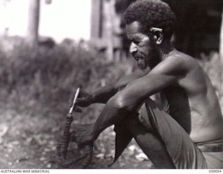 BUKAUA, NEW GUINEA. 1943-10-18. PORTRAIT OF MAHU A VOLUNTEER FOR THE AUSTRALIAN AND NEW GUINEA ADMINISTRATION UNIT LABOUR FORCE, LIGHTING HIS CIGARETTE FROM THE END OF A SMOULDERING COCONUT FIBRE ..